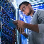 Low angle portrait of young man using digital tablet standing by server cabinet while working with supercomputer in blue light